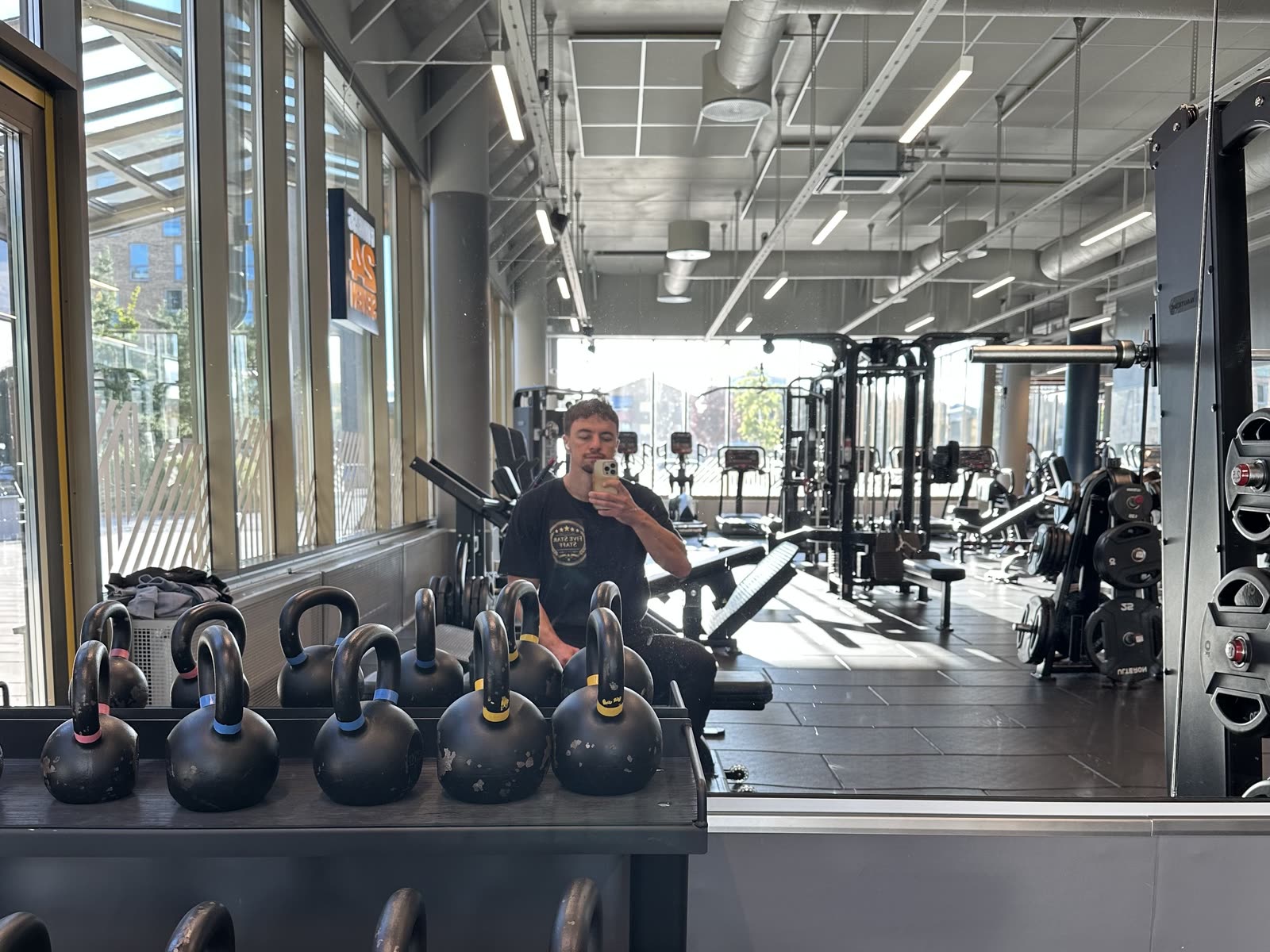 A rack of kettlebells in the foreground of a bright gym, Pelle reflected in the mirror behind them
